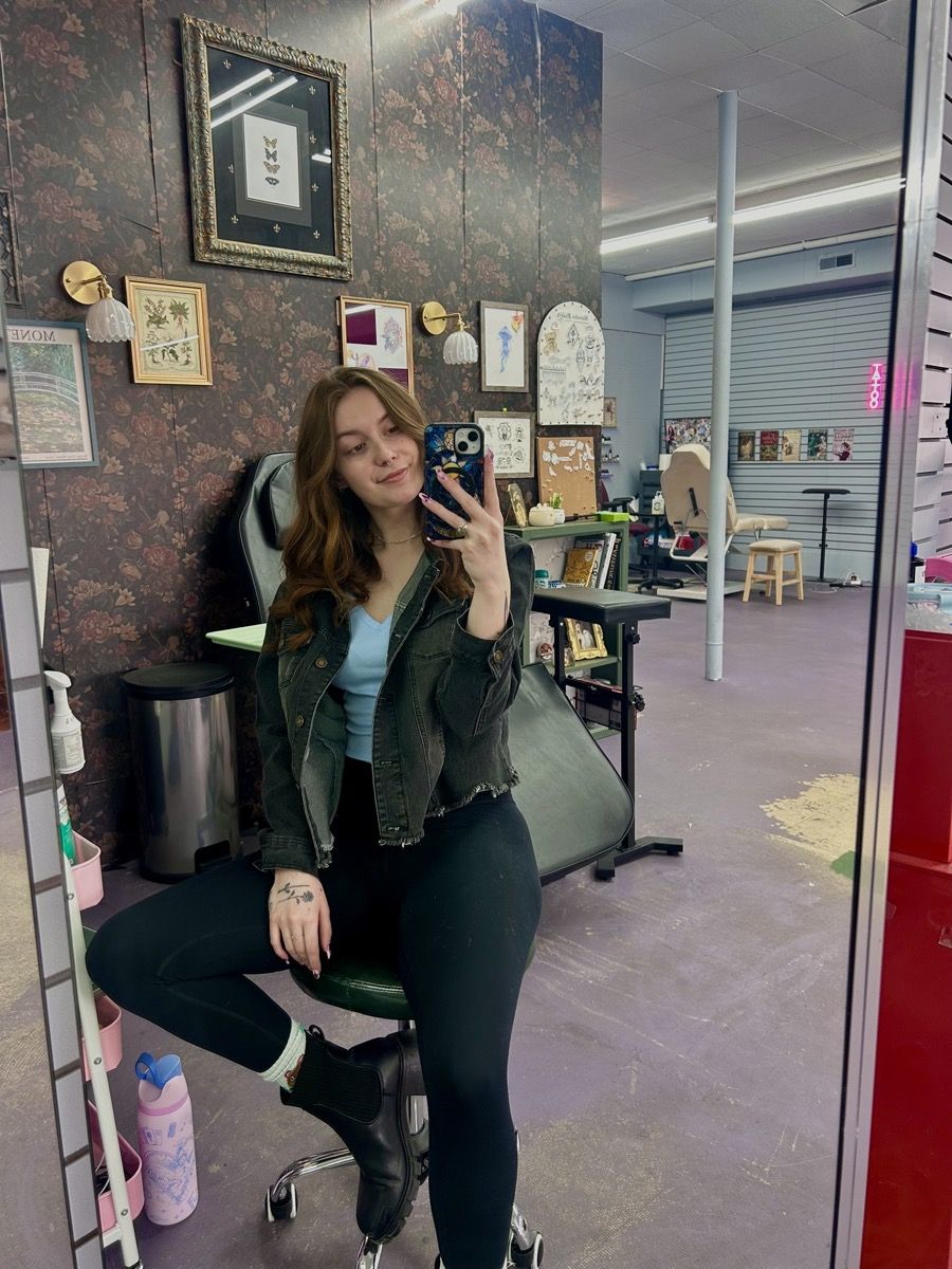 Macy Ella in her booth at Sacred Realm Tattoo Studio, seated in front of a floral wall hung with framed art